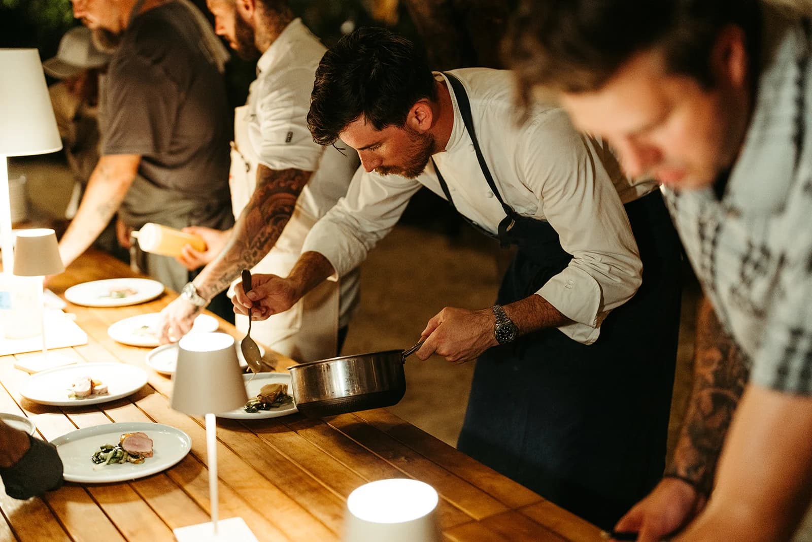 Chefs plating a multi-course dinner by candlelight at a long outdoor table